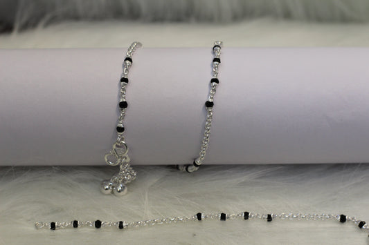 Two silver anklets with black beads, one featuring a small bell-shaped charm, displayed against a white background with a soft texture.
